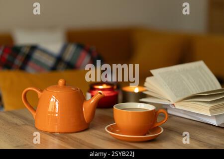 Eine gemütliche Atmosphäre mit einer orangen Teekanne und einer Tasse auf einem Holztisch. Ein offenes Buch und beleuchtete Kerzen schaffen ein warmes, einladendes Ambiente, perfekt zum Entspannen Stockfoto