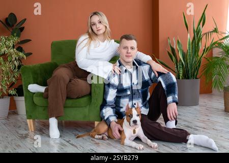 Ein junger Mann und eine junge Frau entspannen sich zu Hause mit ihrem Hund, sitzen auf einem stilvollen Holzboden und Pflanzen in Innenräumen, die den Hintergrund unterstreichen. Stockfoto