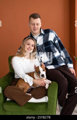 Ein junger kaukasischer Mann und eine junge Frau lächelnd, bequem in einem Sessel mit ihrem weißen und braunen Hund, in einem gut beleuchteten Raum mit orangefarbenen Wänden. Stockfoto