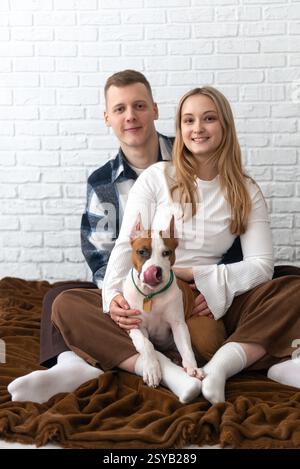 Ein fröhliches junges Paar sitzt zu Hause auf dem Boden und umschließt liebevoll ihren verspielten weißen und braunen Staffordshire Terrier Welpen. Sie sind sm Stockfoto