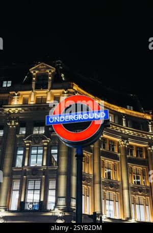 Eine lebendige Aufnahme des klassischen Londoner Underground-Schilds, das nachts vor einer eleganten, beleuchteten Gebäudefassade leuchtet und das Wesen des städtischen Lebens festlegt Stockfoto