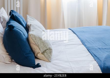 Ein ordentlich gefertigtes Bett mit blauen und beigen Kissen. Die blaue Decke verleiht dem leise beleuchteten Raum einen Farbakzent. Die sanfte Atmosphäre lädt zum Entspannen ein Stockfoto