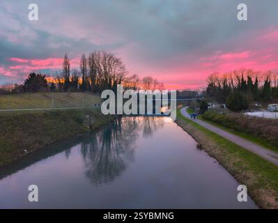 Eine ruhige Kanalszene bei Sonnenuntergang mit leuchtendem rosa und lila Himmel, der sich auf dem Wasser spiegelt. Ein Pfad führt entlang des Kanals, wo zwei Personen laufen. Stockfoto