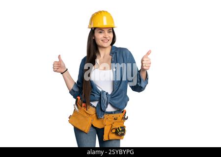 Eine fröhliche Bauarbeiterin mit gelbem Schutzhelm, Denim-Hemd und Werkzeuggürtel zeigt sich doppelt hoch und symbolisiert Selbstvertrauen und Erfolg Stockfoto