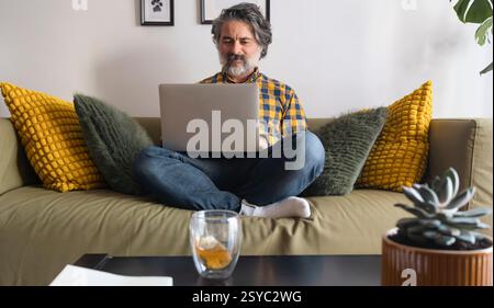 Reifer Mann, der von zu Hause aus auf einem Laptop in einem gemütlichen Wohnzimmer arbeitet Stockfoto
