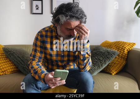 Mann sitzt zu Hause auf dem Sofa und hält ein Smartphone und fühlt sich besorgt Stockfoto