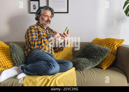 Grauhaariger Freelancer mit Smartphone, der zu Hause auf dem Sofa sitzt Stockfoto