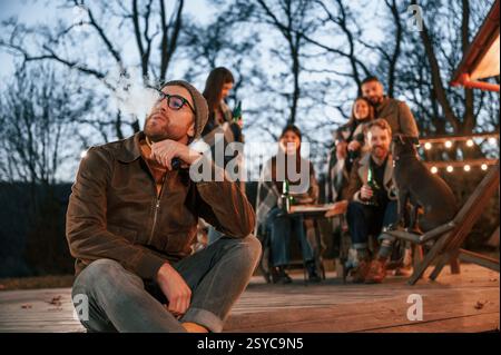 Der Mann raucht eine elektronische Zigarette. Eine Gruppe von Leuten verbringt abends Zeit zusammen auf dem Hinterhof. Stockfoto