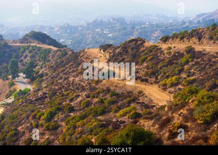 Wanderer navigieren auf dem gewundenen Feldweg im Griffith Park, der eine undurchsichtige Stadtlandschaft mit sanften Hügeln im Hintergrund überblickt. Stockfoto
