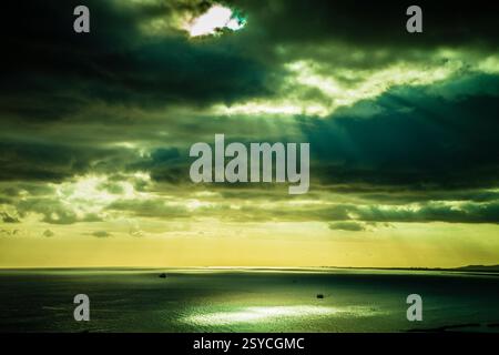 Der Himmel ist grün und bewölkt. Die Sonne scheint durch die Wolken. Es sind Boote im Wasser Stockfoto
