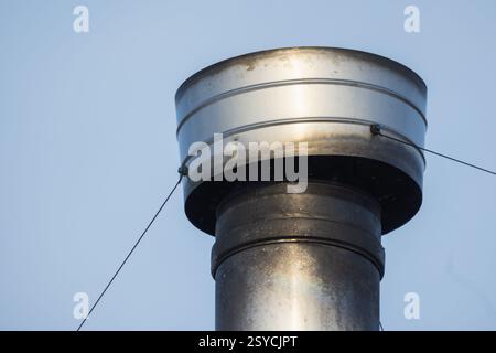 Detaillierte Nahaufnahme eines zylindrischen Metallkaminrohrs mit Schutzkappe unter klarem blauem Himmel, der Wärme aus Industrie und Wohn erfasst Stockfoto