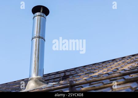 Glänzendes zylindrisches Metallschornsteinrohr mit Schutzkappe befindet sich auf dem Dach des ländlichen Hauses unter klarem blauem Himmel und erfasst Industrie- und Wohnheime Stockfoto