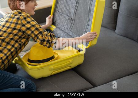 Eine Frau in einem gelben karierten Hemd schließt ihren hellgelben Koffer auf einem grauen Sofa und bereitet sich auf eine Reise vor. Ein stylischer gelber Hut liegt oben auf Stockfoto