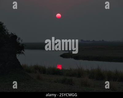 Der Kwando River in Botswana schlängelt sich sanft durch die Landschaft, während die Sonne untergeht und ein warmes, goldenes Leuchten über das ruhige Wasser wirft. Das friedliche Stockfoto