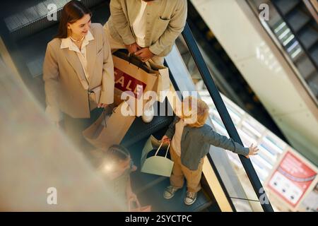 Hochwinkelaufnahme eines blonden Jungen mit Papiertüte, der mit Mom plaudert, während sie eine Rolltreppe benutzen Stockfoto