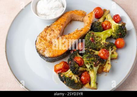 Gebratenes Lachssteak (Forelle) mit gebackenem Brokkoli und Kirschtomaten. Serviert mit Sauerrahmsauce mit Dill. Traditionelle Hausmannskost. Selektiver Fokus, Stockfoto
