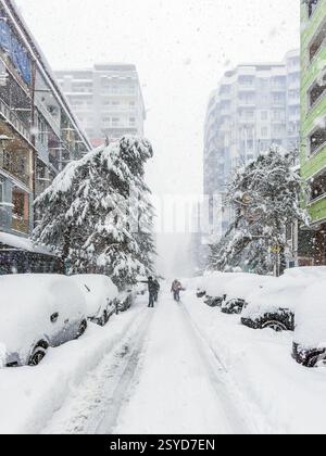 Schneebedeckte Stadtstraße mit geparkten Autos und unter starkem Schneefall vergrabenen Bäumen, umgeben von hohen modernen Gebäuden. Fußgänger, die durch den Wintersturm laufen. Stockfoto