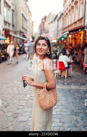 Porträt einer jungen, modischen Frau, die abends in Sommeroutfit auf der Stadtstraße neben dem Restaurant sitzt. Stilvolle Strohhandtasche. Weibliches Zubehör und Tuch Stockfoto
