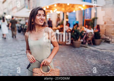 Porträt einer lächelnden, modischen Frau in Sommeroutfit, die in der Abenddämmerung auf der Stadtstraße von Lviv durch Cafés spaziert. Stilvolle Strohtasche und Leinenkleid Stockfoto