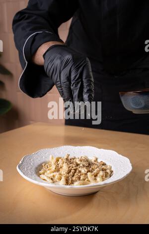 Ein professioneller Koch mit schwarzen Handschuhen garniert einen Teller frischer Pasta mit einem knusprigen Topping und unterstreicht die Gourmet-Präsentation in einem eleganten Re Stockfoto