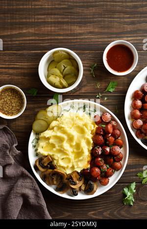 Hausgemachte Kartoffelpüree mit gebratenen süßen und scharfen Würstchen und Pilzen auf weißer Platte auf braunem Holzhintergrund. Draufsicht, flach Stockfoto