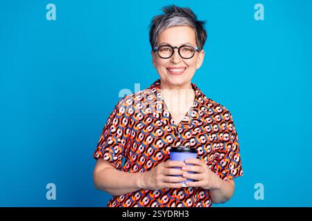 Lächelnde reife Frau in einem bunten gemusterten Shirt hält eine Kaffeetasse vor einem hellen blauen Hintergrund Stockfoto