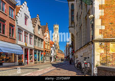 Brügge, Belgien, 5. Juli 2023: Kopfsteinpflasterstraße, alte Gebäudefassade mit Schaufenster und Glockenturm von Brügge Belfort van Brugge Backgr Stockfoto