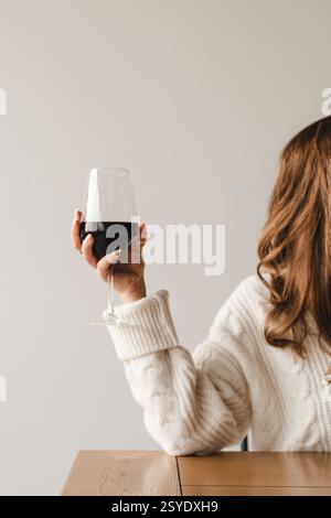 Eine Frau in einem gemütlichen cremefarbenen Pullover hält ein Glas Rotwein an einem Holztisch mit minimalistischer und warmer Ästhetik. Stockfoto