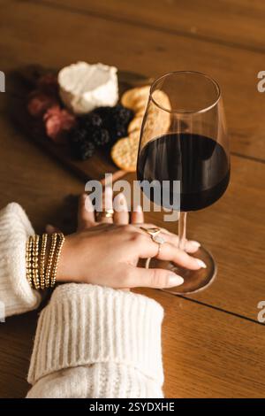 Eine Hand in einem gemütlichen Pullover und Goldschmuck liegt neben einem Glas Rotwein und einer Wurstplatte auf einem Holztisch – elegant und einladend. Stockfoto
