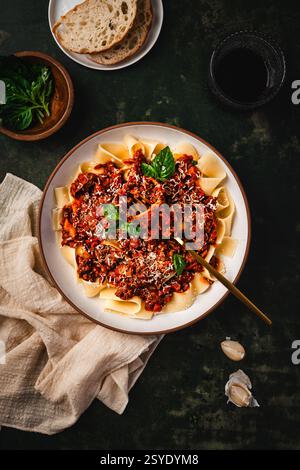 Ein Teller voller Pappardelle Bolognese, belegt mit frischem Basilikum und geriebenem Parmesan, serviert mit rustikalem Brot und Rotwein auf einem dunklen stimmungsvollen Hintergrund. Stockfoto