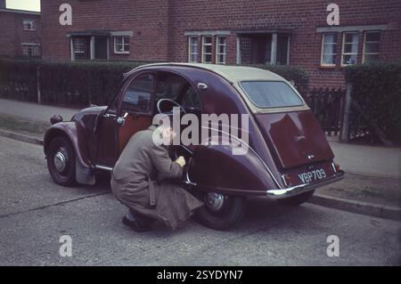 1960er Jahre, historisch, ein junger Mann in einem Regenmantel draußen in einer Straße, kniend neben dem Rücksitz eines kastanienbraunen Citroen 2CV, Nummernschild VBP 709, in einer Straße auf einem Wohngut in England, Großbritannien. Der Wagen, ein Modell AZ, hatte ein durchgehendes Leinendach, eine Wellhaube und Selbstmord-Vordertüren. Der 2CV ist ein klassisches französisches Auto und wird als „People's Car“ bezeichnet. Stockfoto