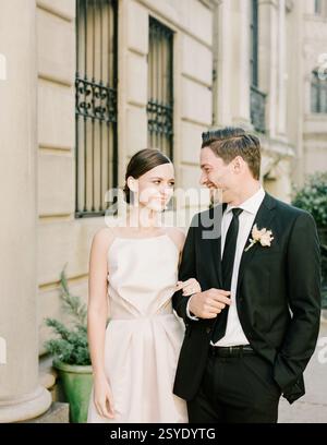 Das Paar teilt einen liebevollen Blick, wenn es formell gekleidet ist, und feiert seine Hochzeit vor einem eleganten Gebäude in einer Stadt. Stockfoto