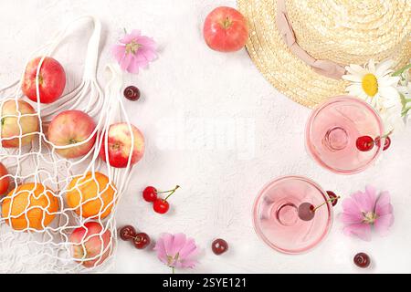 Trendiges Sommerbanner auf hellem Hintergrund. Strohhut, Früchte in Öko-Tasche, erfrischende alkoholische Getränke und Kamillenblüten auf sonnigem Tisch. Urlaub und tr Stockfoto