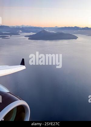 Ein Blick aus einem Flugzeugfenster mit Blick auf die Küsteninseln und Berge bei Sonnenaufgang. Der Flugzeugflügel und das Triebwerk stehen im Kontrast zur riesigen Meereslandschaft darunter. Stockfoto
