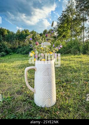Ein weißer Keramikkkrug gefüllt mit bunten Wildblumen auf einem grasbewachsenen Feld mit Bäumen im Hintergrund. Eine natürliche und rustikale Outdoor-Szene. Stockfoto