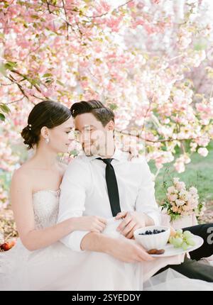 Ein Paar sitzt eng zusammen unter Kirschblüten und teilt ein leichtes Picknick inmitten rosafarbener Blüten und Grün. Stockfoto
