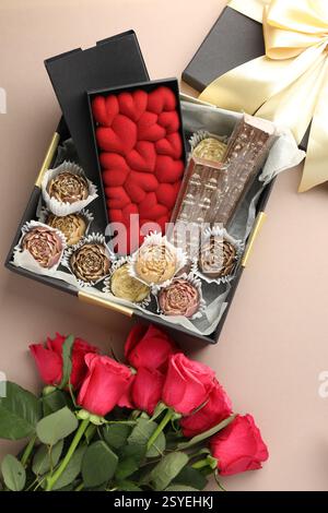 Verschiedene köstliche Schokoladenbonbons in der Box und Rosen auf beigefarbenem Hintergrund, Blick von oben Stockfoto