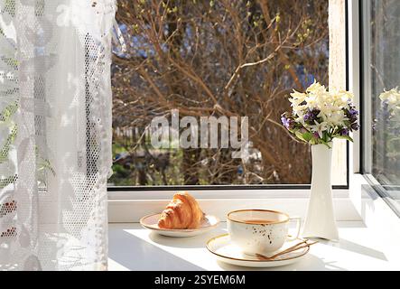 Tasse Tee, Croissant und Frühlingsblumen in einer Vase an einem sonnigen Fenster. Das Konzept des Wohnkomforts und der Begrüßung des Frühlings. Wunderschönes Blumenarrangement mit Stockfoto