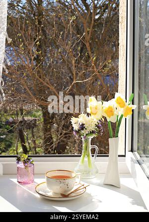 Tasse Tee, Croissant und Frühlingsblumen in einer Vase an einem sonnigen Fenster. Das Konzept des Wohnkomforts und der Begrüßung des Frühlings. Wunderschönes Blumenarrangement mit Stockfoto