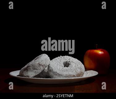 Eine Nahaufnahme von Donuts in Pulverform auf einer Keramikplatte, begleitet von einem roten Apfel, vor dunklem Hintergrund, wodurch eine stimmungsvolle Komposition entsteht. Stockfoto