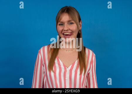 Eine verspielte junge Frau, die ihre Zunge in einem Studio mit blauem Hintergrund ausstreckt Stockfoto