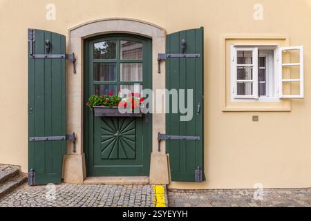 Die charmante Hausfassade verfügt über eine lebendige grüne Tür mit dekorativen Schnitzereien, passenden Fensterläden, einem Fensterkasten mit roten Blumen und einem angrenzenden weiß Stockfoto