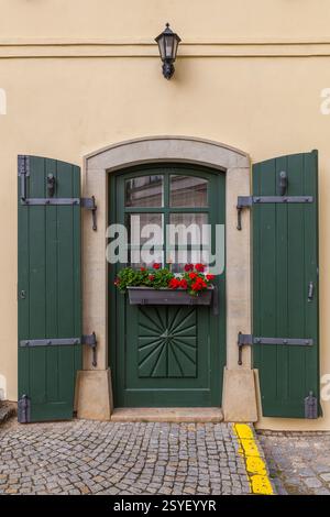 Die charmante Hausfassade verfügt über eine lebendige grüne Tür mit dekorativen Schnitzereien, passenden Fensterläden, einem Fensterkasten mit roten Blumen und einem angrenzenden weiß Stockfoto