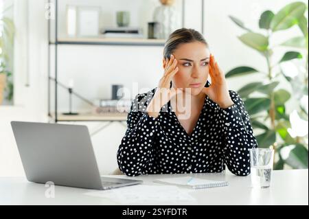 Eine verärgerte, müde Geschäftsfrau, die sich die Schläfen reibt, Kopfschmerzen erleidet, während sie in einem modernen Büro mit einem Laptop an ihrem Schreibtisch sitzt, besorgt und tief in Gedanken aussieht, braucht eine Pause von der Arbeit Stockfoto