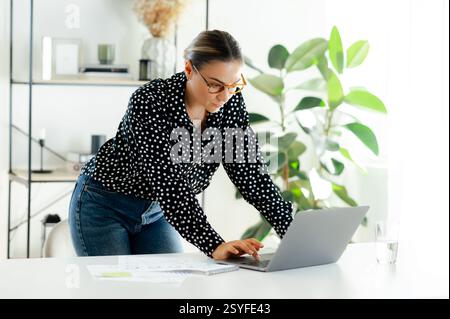 Konzentrierte kaukasische Geschäftsfrau mit Brille, Firmenchef, die am Schreibtisch in einem modernen Büro steht, an einem Laptop arbeitet und Dokumente mit einem ernsten Ausdruck liest Stockfoto