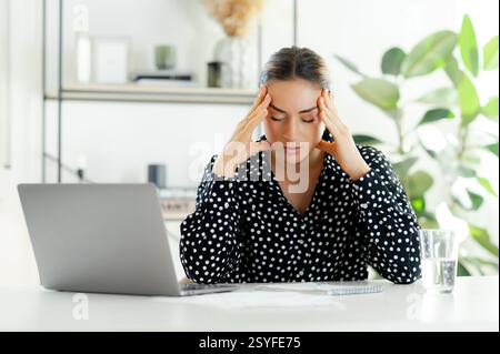 Gestresste, erschöpfte Geschäftsfrau mit geschlossenen Augen, sitzt an einem Schreibtisch mit Laptop, reibt sich mit beiden Händen an den Schläfen, überfordert und müde im modernen Büro, fühlt Kopfschmerzen und überarbeitet, braucht Ruhe Stockfoto