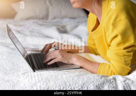 Frau in gelbem Pullover, die auf dem Bett liegt und an einem Laptop arbeitet Stockfoto