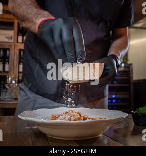 Ein professioneller Koch in schwarzen Handschuhen garniert frisch zubereitete Pasta mit geriebenem Käse in einem feinen Restaurant. Die Szene fängt ein elegantes f Stockfoto