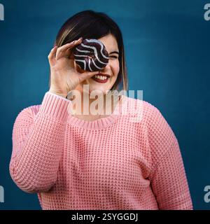 Fröhliche junge Frau in einem pinkfarbenen Pullover, die verspielt einen Schokoladenkrapfen mit Glasur über dem Auge hält und vor einem leuchtend blauen Hintergrund lächelt. Spaß und Stockfoto