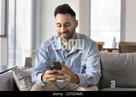 Hübscher hispanischer junger Mann aus den 30er Jahren, der ein Smartphone benutzt, das auf dem Sofa sitzt Stockfoto